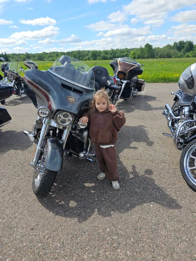 A young child lies prone on a large motorcycle parked on an asphalt surface, with additional motorcycles visible in the background. The child wears a brown tracksuit and light-colored sneakers, and her hair is blonde. The motorcycle beneath her is a dark gray color with chrome accents. A field and treeline appear in the background under a partly cloudy sky. The scene appears to be outdoors, possibly at a motorcycle event or gathering. There is no evidence to confirm it is in the city of Borodinskogo Muzeya, Russia.
