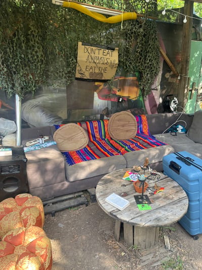 The image depicts an outdoor or semi-enclosed living area located in the city of Sint-Pieters-Leeuw, Belgium. The scene features a grey L-shaped sectional sofa positioned centrally, adorned with two vibrantly colored, horizontally striped blankets in shades of red, blue, green, yellow, and orange, along with two circular, light brown button-tufted cushions. In front of the sofa stands a round coffee table fashioned from a large wooden cable spool. An open book with white pages rests on the table, alongside a dark, multi-tiered decorative stand holding various small objects, a small terracotta pot, scattered coins, a small green sign, and a small, colorful, possibly rainbow-striped item.
To the left of the sofa, a dark speaker unit is situated on the ground, with a small white musical keyboard and a clear spray bottle on the sofa's armrest. To the right of the sofa, a light blue hard-shell suitcase with black accents is visible, along with a black power cord reel. The ground is exposed earth.
Overhead, green camouflage netting hangs, interspersed with a yellow corrugated pipe and what appears to be string lights. Behind the sofa, a large mural or poster depicting industrial or mechanical elements in warm tones of red, orange, brown, and grey serves as a backdrop. A large grey bag or sack is present on the far left. A prominent cardboard sign hangs above the sofa, secured with black tape, displaying the hand-written text "DON'T EAT ANIMALS" above "EAT THE" followed by a crude drawing resembling human brains, a dollar sign symbol, and a pig's snout or footprint. No individuals are present in the scene, and no actions or interactions are depicted.