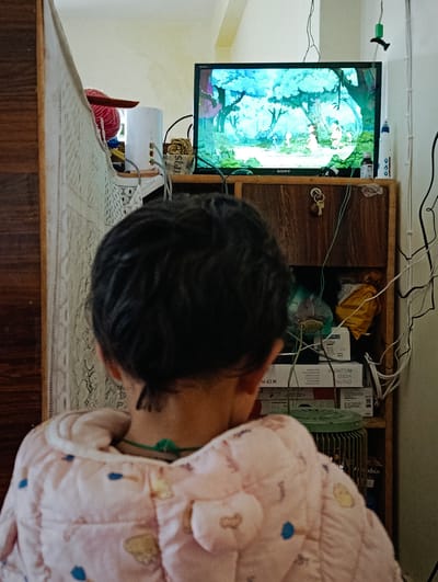 The media file shows a rear view of a young child wearing a pink quilted jacket with blue and yellow embellishments. The child is facing a television screen which displays an animated scene featuring trees and figures. The television is on a wooden cabinet, which is cluttered with various objects including wires, boxes, and what appear to be craft supplies like yarn. A set of keys hangs from the cabinet door. The setting appears to be indoors, likely a home, given the domestic nature of the objects and the presence of a television. The lighting suggests it could be daytime, though the exact time is not discernible. There is no indication of weather.
Notable details include the brand name "SONY" visible on the television. Text on a box beneath the television reads "DIGITAL VIDEO RECORDER". Other boxes and items are partially obscured. A white cylindrical object and a red ball of yarn are visible on the cabinet shelf to the left of the TV. The child's dark hair is visible, and they are wearing a green necklace. The overall scene conveys a sense of quiet engagement, with the child focused on the entertainment provided by the television. The location context provided is Gemur, India.