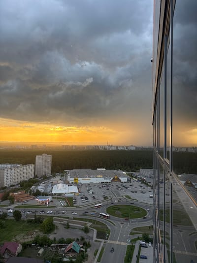 The image is a high-angle, full shot of a city scene in Boldino, Russia, at dusk. The sky is filled with dramatic, dark gray clouds, with a sliver of bright yellow-orange sunset breaking through on the left horizon. A large, modern glass-covered building dominates the right side of the frame, reflecting the sky and surrounding cityscape. In the middle ground, a large, light-colored retail building with yellow signage is visible, along with a vast parking lot filled with numerous cars. A circular intersection with a roundabout is prominent, with a red and white bus transiting it. Further back, dense green forest stretches across the landscape, behind which lie rows of apartment buildings. In the foreground, on the left, are a few smaller residential buildings and a glimpse of a green area. The overall impression is a view from a high-rise building overlooking a commercial and residential district under a stormy, yet vibrant, sky.