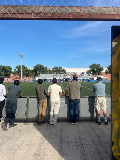 The image shows a view through a chain-link fence onto a soccer field in Sant Adrià de Besòs, Spain. Several men are standing at the fence, watching a soccer game. The field is artificial turf, and there are spectators in the bleachers in the background. The weather is sunny and clear. The men in the foreground are dressed casually, some in jeans and t-shirts, others in shorts and t-shirts. The fence is made of metal and has a rusty top railing. There is a large yellow pole with graffiti on the right side of the image.