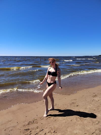 A young woman with red hair and sunglasses stands on a sandy beach in Solnechnoye, Russia, wearing a black bikini. She has tattoos on her left arm. The sun is shining brightly in a clear blue sky, and gentle waves lap at the shore of a large body of water, likely the Gulf of Finland. In the distance, several people can be seen swimming or wading in the water. The sand is wet near the water's edge, and footprints are visible in the drier sand. The woman's shadow is cast long on the sand behind her.