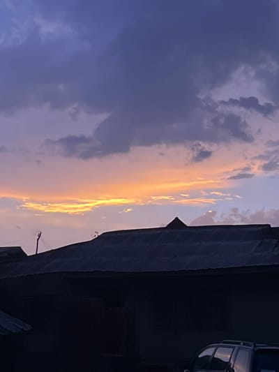 The image shows a dusky sky with orange and purple-grey clouds. A building with a corrugated iron roof occupies the right side of the frame. A vehicle is partially visible on the right edge, near the upper corner of the frame. A pole with a wire is visible in front of the roof. The location is likely Iseyin, Nigeria, based on the prompt.