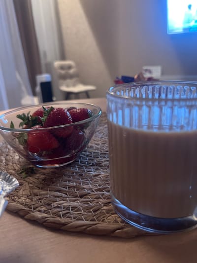 In Yastrebovskii, Russia, on a table covered with a woven placemat, a glass bowl filled with ripe strawberries sits next to a tall glass containing a light brown beverage. The background is dimly lit, with hints of a television screen displaying a bright image and out-of-focus furniture, suggesting an indoor setting. The overall scene depicts a simple, casual moment, possibly a snack or a light refreshment.