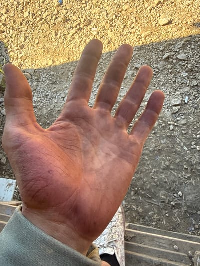 A close-up, over-the-shoulder view of a dirt-covered human hand is presented, palm up and fingers extended, against a background of gravel and dirt. The skin on the palm and fingers is reddish-brown with visible dirt ingrained in the creases. The fingernails also appear dirty. The person's forearm, covered by a light green sleeve, is visible at the bottom left. A portion of a white sneaker with light gray laces is visible at the bottom right, suggesting the person is standing. The environment appears to be outdoors, with a textured ground composed of small, light-colored stones and darker soil with debris. There are no identifiable landmarks or features that would indicate a specific location within Matchino, Russia.