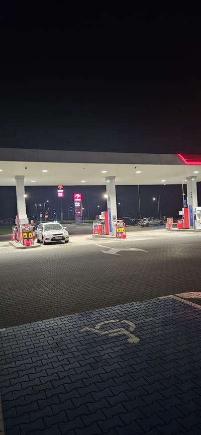 The image shows a night view of a gas station in Sobieszczany-Kolonia, Poland. The gas station has multiple pumps under a canopy lit by fluorescent lights. There are several cars at the station, including a white car with its driver standing next to it at one of the pumps. Two large "ORLEN" signs with "stop.cafe" are visible in the background. The ground is paved with interlocking bricks. In the foreground, a white symbol indicating a disabled parking spot is painted on the pavement. The overall scene is well-lit by the gas station's artificial lighting, contrasting with the dark night sky.