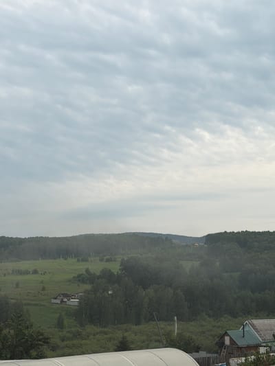 The image depicts a rural landscape under a cloudy sky. In the mid-ground, a cluster of buildings, including a house with a dark roof and light-colored walls, is visible surrounded by a white fence, situated on a grassy hillside. Further into the distance, rolling hills covered in dense green trees dominate the horizon. Some scattered, smaller structures and utility poles are also discernible in the background. The foreground shows the edge of a structure with a curved, white, translucent roof, partially obscuring the view of the landscape beyond. Another building with a green roof and a visible ladder is in the lower right corner. The overall scene suggests a quiet, overcast day in a rural area, likely in the vicinity of Kuznetsovo, Russia, given the context.
