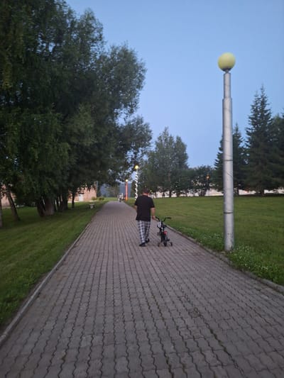 A person is walking along a paved walkway in a park-like setting. The person is wearing a black t-shirt and checkered pants. They are pushing a small black bicycle. The walkway is made of interlocking grey pavers and is lined with grass on one side and trees on the other. There are several trees of varying sizes, some appearing to be deciduous and others coniferous. In the background, there are buildings and more trees. A tall lamppost with a yellow globe on top is visible to the right of the walkway. The sky is a clear, pale blue, suggesting it is either early morning or late evening. The scene appears to be in a residential area or a park in the city of Atamanovka, Russia.