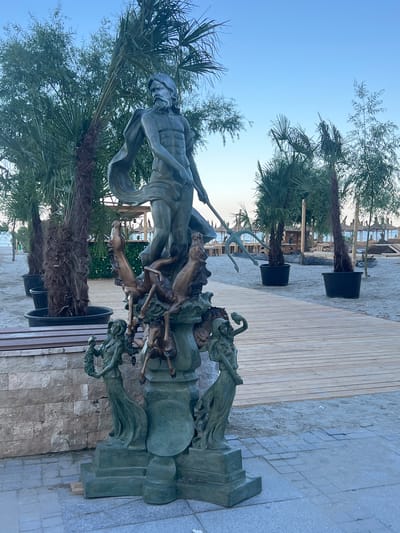 The image depicts an outdoor scene, likely in the city of Năvodari, Romania, featuring a bronze statue prominently in the foreground. The statue portrays a male figure, possibly Neptune, holding a trident and surrounded by horses and female figures at its base. The statue is situated on a tiled area adjacent to a sandy beach. Palm trees in large planters are visible behind the statue, and a wooden pathway leads further into the beach area. In the background, beach umbrellas, lounge chairs, and some structures suggest a resort or public beach setting. The sky is clear and pale blue, indicating either early morning or late evening. The overall environment is that of a seaside location, possibly a promenade or entrance to a beach. There are no people visibly interacting with the statue or in the immediate vicinity. The scene is static and captures a moment of stillness.