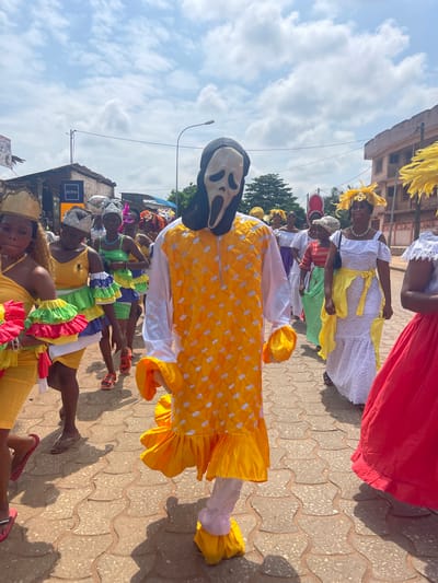 This is a street-level, eye-level shot taken outdoors in Porto-Novo, Benin, under a partly cloudy sky. A procession is underway, with several individuals in colorful attire. In the foreground, a person is wearing a white and yellow costume with a mask resembling the character Ghostface from the Scream movie franchise. This costumed individual is facing the camera and appears to be walking forward. Behind this person, a group of women is also participating in the procession, many of them wearing brightly colored dresses and elaborate headwear. To the left, a woman in a yellow dress with ruffled sleeves is visible. To the right, a woman in a white lace dress with a yellow sash is walking. The ground is paved with interlocking bricks. In the background, there are buildings, some appearing commercial with signage, and streetlights. The overall atmosphere suggests a festive or celebratory event.
