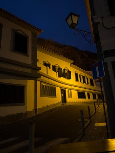 An upward-angle nighttime shot shows the lit facade of a building next to a street in Ponta do Sol, Portugal. A blue-and-white "Dead End" sign is near the top of the frame. The architecture features a light yellow, tall building with dark windows and window coverings. The facade is illuminated, providing contrast to the dark night. A wrought-iron street lamp with a glowing rectangular bulb is also visible. Adjacent to the building is a dark, narrow street and parts of a terraced hill. The sky is deep blue.