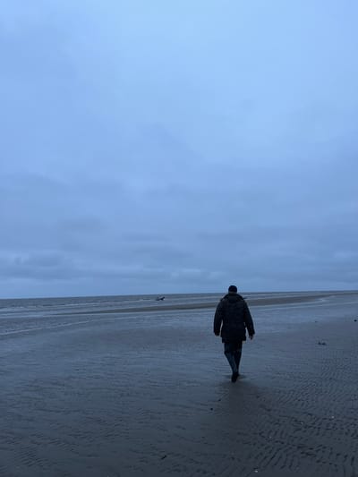 A person walks along a wet sandy beach with the ocean in the background under a cloudy sky. The person is wearing a camouflage jacket and dark pants, facing away from the camera. In the distance, a small inflatable boat is partially submerged in the water. The beach is marked with ripples from receding water. The location is Yandova Guba, Russia.