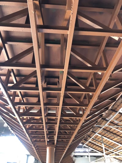 The image shows a close-up, low-angle view of the underside of a building's roof structure, composed of numerous brown, painted metal beams. These beams form a complex lattice of intersecting supports, including horizontal beams, diagonal braces, and vertical struts. The metal appears to be steel, with visible rivets and joins. Above the metal framework, a brown, opaque material forms the underside of the roof. To the right of the image, a portion of a thatched roof structure is visible, supported by white metal beams. Natural light filters through gaps in the structure and from the left side of the image, creating shadows. The scene is industrial or construction-related. This image was taken in Uteungeulinggang, Indonesia.