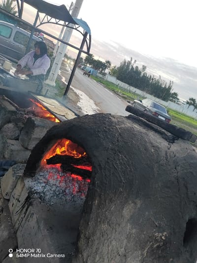 The image captures an outdoor scene featuring traditional cooking activities, likely selling freshly made flatbreads by the roadside.

In the foreground on the right, a large, dark, dome-shaped oven, possibly made of clay or mud (a *tabun* or similar), is prominently featured. Its arched opening reveals bright orange flames and glowing red embers, indicating it is actively heating or in use. Several charred logs are visible within the firebox.

To the left of the oven, slightly further back in the mid-ground, a woman is engaged in preparing food. She wears a light-colored headscarf, a pale purple or white patterned sweater, and patterned pants. She stands beneath a rustic metal canopy structure with a blue fabric or tarp covering part of its roof. In front of her is a flat metal cooking surface (griddle or *saj*) placed directly over an open fire. Smoke or steam rises from this area, and several flat, round dough pieces, resembling flatbreads, are visible cooking on the hot surface. The woman's hands are positioned over the griddle, suggesting she is either flattening dough or tending to the cooking bread.

The background reveals a street or road running diagonally through the frame from the mid-left towards the upper right. On the left side of the road, a light-colored van is visible, partially obscured by the canopy structure. Further down the road, a small, light-colored cart or wagon can be seen. On the right side of the road, a dark-colored sedan (possibly an older model Mercedes-Benz) is parked, facing away from the viewer. The road is bordered by a grassy verge and a fence, behind which there are rows of green trees and bushes, including some palm trees. The sky is bright but overcast, suggesting either early morning or late afternoon light, creating a soft, diffused illumination across the scene.

The ground around the cooking areas appears to be dirt or earth, with some patches of lighter-colored, possibly wet, ground near the road.

A digital watermark is visible in the bottom left corner of the image, stating "HONOR 70" and "54MP Matrix Camera".