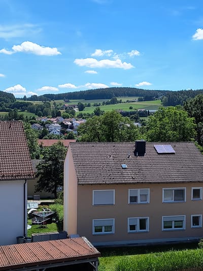 The image depicts an aerial view of a rural landscape in Oberviechtach, Germany. The scene is dominated by a bright blue sky filled with scattered white clouds, suggesting fair weather and daytime. On the right side of the frame, a modern, multi-story building with a beige facade and numerous windows is prominent. The building has a tiled roof, and on one section of the roof, two solar panels are visible, along with a chimney.
Below the building, a well-maintained green lawn is visible, with garden furniture, including a clothesline and chairs, set up on it. Further to the left, the landscape transitions into a dense, dark green forest that stretches across the horizon. Interspersed within the forest and in the foreground are scattered houses with red-tiled roofs, forming a small village. The houses are surrounded by lush green trees, indicating a season of growth, likely summer. The rolling hills and fields suggest a pastoral setting. The image is taken from a high vantage point, possibly from a drone or a very tall structure, looking downwards and across the landscape. There is no visible text in the image. The overall impression is one of a peaceful, sunny day in a picturesque German countryside.