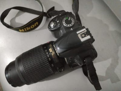 The image shows a Nikon DSLR camera with a telephoto lens attached. The camera has a black body and a textured grip. A Nikon branded camera strap, with the logo clearly visible in yellow text on a black background, is draped around the camera. The lens has markings for focal length, including 200mm, 135mm, 105mm, 85mm, 70, and 55. The camera is placed on a light-colored, textured surface, possibly a table or desk. The lighting suggests an indoor setting, with shadows cast on the surface behind the camera. No people or specific location cues are visible beyond the implied indoor setting and the camera brand. The image appears to be a product shot or a personal photo of camera equipment.