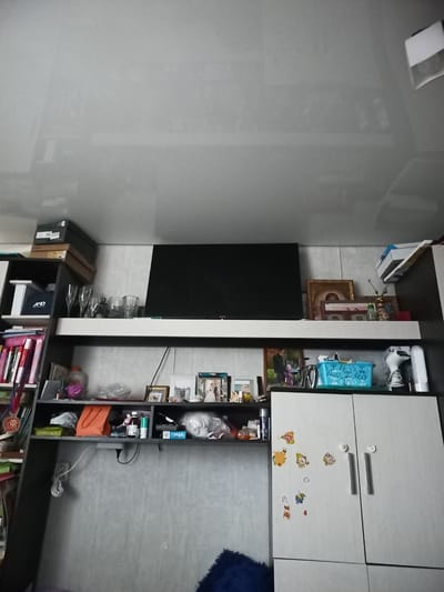 A shelf unit with a television is displayed in an indoor setting. The shelf unit is dark brown and gray. A large, black television is mounted on the wall above the shelves. The shelves themselves are filled with various items, including books, boxes, glassware, and framed pictures. On the top shelf, to the left, are stacks of boxes. Below that, on a shelf to the left, is a black tote bag with "AND" written on it. Several tall glasses are on the shelf next to the tote bag. Further down, a collection of books is visible on the left. On the shelf below the television, there are framed photos, small decorative items, and a blue basket. To the right of the shelf unit, a white cabinet with two doors is visible. The cabinet is decorated with several colorful stickers of cartoon characters. The overall impression is of a living space or entertainment area. The image appears to be taken in Lomonosov, Russia, based on the context provided.