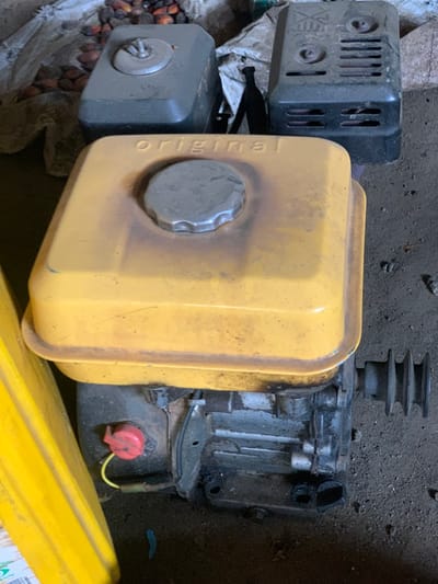 The image shows a small gasoline engine, likely for a generator or other small machinery, lying on the ground. The engine has a yellow fuel tank with the word "ORIGINAL" written on the side. The engine is partially covered by a black plastic cover. The engine is lying on a rough surface that appears to be dirt or gravel. There is a red switch on the engine. The coordinates provided, latitude: 7.12, longitude: 5.362, place the image in the Egbeda area of Lagos, Nigeria. The engine is in a state of disrepair and is covered in dirt and grime. There is no one in the scene. There is a yellow plastic container in the bottom right of the image. The image is likely taken from above, at a slightly tilted angle. There are other objects visible in the image, including a pile of what appears to be palm fruit, but they are not in focus.