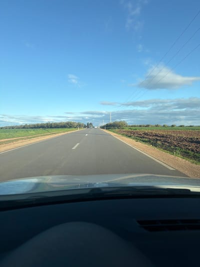 The scene depicts a straight, two-lane asphalt road, likely within or near Sidi Slimane Moul Al Kifane, Morocco, viewed from inside a vehicle looking forward through the windshield. The vehicle's dashboard and a reflection are visible in the lower foreground. The road features a solid white line on the right shoulder and a dashed white center line, which transitions to a solid line further down, indicating a two-way thoroughfare.
To the left of the road, green agricultural fields extend, bordered by a dense line of trees or shrubs in the mid-ground. To the right, the immediate roadside consists of a gravel shoulder transitioning into tilled brown earth and some green vegetation. A series of wooden utility poles carrying overhead power lines run along the right side of the road, receding into the distance alongside sparse trees.
Approximately three light-colored passenger vehicles are visible in the far distance, traveling away from the camera in the same direction. The sky is clear blue with scattered white and grey clouds, particularly towards the horizon. The environment is rural, illuminated by direct sunlight, indicating daytime. No individuals are explicitly visible.