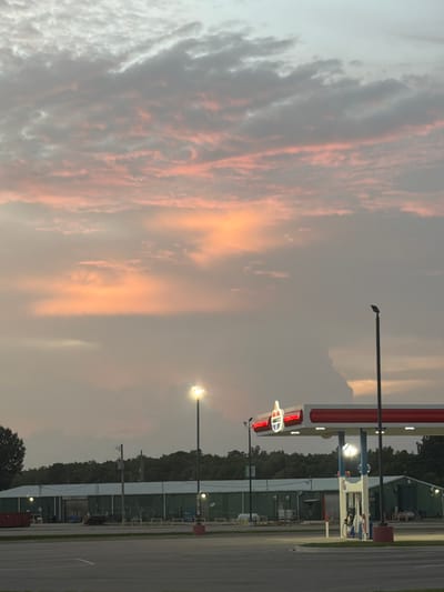 The image shows a gas station and a building in Cardwell, United States, during sunset. The sky is filled with colorful clouds, ranging from pink and orange to gray. A gas station with an Amoco sign is prominently featured in the foreground, with a few gas pumps visible. A long, green building with a white roof is situated behind the gas station. The scene appears to be tranquil, with no people or vehicles present. The overall atmosphere is serene, with the warm hues of the sunset creating a calming effect.