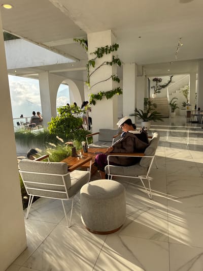 The image captures an interior scene in Kedisan, Indonesia. An individual wearing a head covering and a dark coat is seated in a light-gray chair with white metal legs. The chair is positioned at a wooden table. Next to the chair is a round, gray ottoman. Other people are seated at the table, surrounded by decorative plants and foliage. The background features an open-air architecture with white walls, archways, and views of the sky. The floor is tiled with a white marble pattern, and sunlight is cast upon it. The setting appears to be a modern, open-air cafe or lounge.