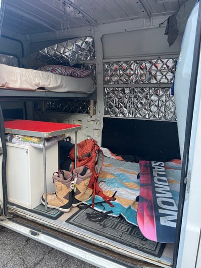 This image displays the interior of a white cargo van, located in Burgstall, Austria, converted for living and travel. On the left side, a wooden-framed bunk bed system is present; the upper bunk features white bedding with a red and white patterned pillow, and the lower bunk is also fitted with white bedding. Below the lower bunk, a white, rectangular storage cabinet stands next to a portable table with a red tabletop and metallic support legs. Small decorative lights are strung along the ceiling above the bunks. The van's upper interior walls and ceiling are lined with silver, diamond-patterned thermal insulation material. On the floor, near the open side door, a pair of tan and black snowboard boots rests on a dark, patterned floor mat. Adjacent to the boots, an orange hiking backpack is visible. Further to the right, a black and pink Salomon brand snowboard lies flat on a light-colored, striped sleeping pad that covers a portion of the floor. The arrangement indicates a setup for cold-weather recreational activities.
