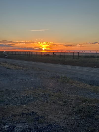This media file captures a sunset scene in Bozshakol, Kazakhstan. The image is oriented vertically, presenting a landscape divided into distinct zones. On the left, the sky is a gradient of soft blues and oranges, illuminated by the setting sun peeking through scattered clouds. The sun itself is a bright, fiery orb, casting a warm glow that bathes the clouds in vibrant hues of orange and yellow. A tall, imposing fence with barbed wire runs horizontally across this section, marking a boundary between the sky and the land beyond.
To the right of the fence, a dirt road stretches diagonally across the frame, appearing worn and textured. The road is bordered by dry grass and dark, rocky terrain. The colors in this area are muted, dominated by grays, browns, and dark greens, contrasting with the vivid sky. A small, dark object, possibly a signpost with a camera or sensor, is visible near the fence and road. The overall impression is one of a remote, perhaps industrial or security-related location at the end of the day. The time of day is clearly sunset, with the warm, dramatic lighting indicating this. The weather appears clear with some cloud cover. No people are visible in the image. No discernible text is present.