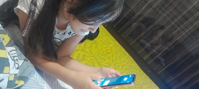 A medium shot captures a young person with dark hair, wearing a white t-shirt, engrossed in their smartphone. The person is positioned on a bed with a patterned duvet featuring geometric shapes in white, yellow, and blue. To the right of the bed, a textured, bright yellow surface is visible, possibly a carpet. Behind this, a dark wooden paneling with a distinct grain pattern creates a backdrop. The person's hands are holding the phone, which displays a bright blue screen with white icons. The lighting suggests an indoor setting. The image is taken from a slightly high angle, looking down at the subject. The overall scene appears to be within a private residence.