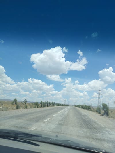 The image shows a paved road in the city of Besapan, Uzbekistan, under a clear blue sky with scattered white clouds. The road appears to be in moderate condition with some wear and tear. The surrounding landscape is arid with sparse vegetation, consisting of small trees and shrubs. Power lines and transmission towers are visible in the distance, indicating the presence of infrastructure. The perspective suggests the photo was taken from inside a vehicle, looking forward along the road. No people or specific actions are depicted. The overall scene is one of a rural or semi-rural environment with a sense of open space.