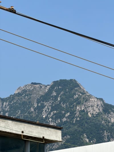The image shows a mountainous landscape with power lines in the foreground. A building is partially visible in the lower left corner. The mountain is covered in trees and rocks, and the sky is clear blue. The scene appears to be from a city or town at the base of the mountains, possibly with industrial or urban infrastructure.