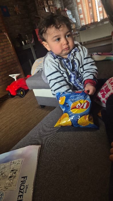 An infant or toddler, with dark curly hair and a light complexion, is positioned on a grey upholstered sofa. The child wears a blue and white plaid shirt underneath a grey and black striped cardigan, featuring a red cuff on the right wrist. The child's right hand holds a partially open blue bag of Lay's Paprika Flavour potato chips, with several chips visible both within the bag and spilled onto the dark grey fabric surface of what appears to be an adult's lap or an adjacent cushion. The child's gaze is directed towards the upper right, exhibiting a neutral expression. In the lower left foreground, a book cover displaying "Disney Frozen" is partially visible on the dark grey upholstered surface. To the left of the child, on a light brown carpeted floor, a red toy push-car with a white steering wheel is present. The background features a brick-patterned wall with multiple framed pictures mounted upon it. Further in the background, a window covered by vertical orange/brown blinds is visible. A dark surface with metallic objects, possibly containers, is also present on the left side of the background. The scene depicts an indoor residential setting. The image itself does not contain identifiable landmarks or specific geographic indicators to independently confirm the location as Melle, Belgium.