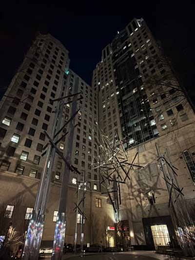 Two tall, beige buildings at night are angled in the image. A complex metal art structure is positioned in the center, casting shadows on the building façades. Many windows are visible, with some illuminated. Part of a building with a dark window and faint red signage appears in the background. The sky is dark.
