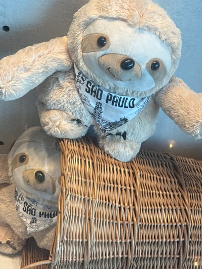 The image captures two light brown plush sloth toys displayed within an indoor retail or souvenir setting in Guarulhos, Brazil. The upper sloth, positioned centrally, rests its lower body on the rim of a prominent, light brown woven wicker basket. This sloth faces slightly right, directly towards the viewer, and wears a white bandana with "SAO PAULO" printed in black capital letters, accompanied by line art depicting city landmarks. Its eyes and nose are black, glossy plastic components. A second, identical sloth is partially visible in the lower-left quadrant, peeking from behind the wicker basket, also wearing a "SAO PAULO" bandana. The background is a plain, light-toned wall or surface, exhibiting subtle reflections from ambient lighting. The wicker basket forms a structured, textured base for the display.