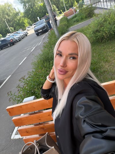The image shows a woman with blonde hair, wearing a black jacket, sitting on a wooden bench in what appears to be an outdoor urban environment. There's a parking lot with multiple vehicles visible in the background. The location is identified as Grebeni, Russia. A light-colored purse is visible beside the bench. The woman is looking at the camera, posing for a selfie. There are trees and greenery in the background, suggesting a park or green space near the parking area. There is a bracelet visible on the woman's wrist.