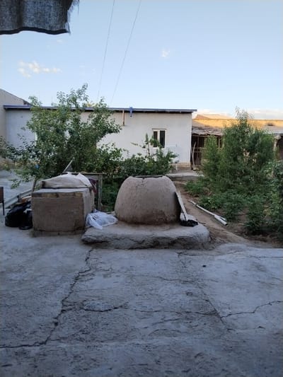 The image depicts an outdoor courtyard with a tanpyr oven in the foreground, a common feature in Central Asian cuisine, particularly in places like Chalysh, Uzbekistan. The tanpyr, a conical clay oven, sits on a raised stone platform. To its left is a rectangular clay structure, possibly for storage or preparation, covered with a white cloth. A black plastic bag and a white plastic bag are visible on the ground near these structures.
Behind these elements, a light-colored building with a dark roof trim and a single window is visible. Trees with dense green foliage are planted around the building and in the background. The ground in the courtyard is paved with concrete, showing visible cracks. The sky above is a light blue with a few scattered white clouds. The overall scene suggests a rural or semi-rural setting, likely a residential area in Chalysh.