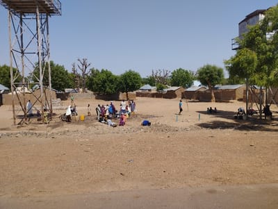 The image shows a scene in Tongo, Nigeria, with a large water tower in the foreground and a smaller water tower in the background. Several people, mostly children, are gathered in a dirt area in front of the larger water tower. Some are sitting on the ground, while others are standing. There is a motorbike in the background. The scene appears to be a gathering place for the community. The ground is dry and sandy, and there are several trees in the background. The houses in the background are simple, made of mud bricks. The sky is clear and blue.