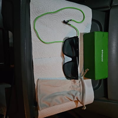 The image shows a pair of black Polaroid sunglasses with a green cord attached to the temples, a green Polaroid box, and a grey drawstring pouch. These items are resting on a white paper towel which sits on a black dashboard in the interior of a car. There are no people visible. The location is not identifiable as Vic, Spain based on the image.