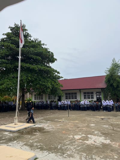 In Langnibong, Indonesia, a school gathering is underway on an overcast day. A group of students, predominantly dressed in gray uniforms and white headscarves, stand in formation near a light green building with a red tiled roof. Some students are in a line, while others are dispersed. A flag pole with the Indonesian flag, red and white, stands prominently to the left, partially obscured by a large, leafy tree. Two boys are walking across the wet, muddy courtyard in the foreground, one carrying a backpack. The environment is a schoolyard with puddles on the ground, indicating recent rain.