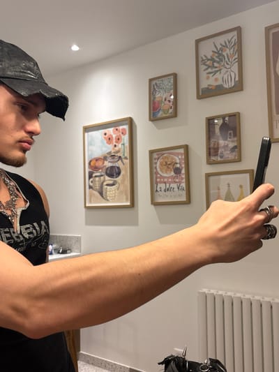 The scene is an interior location in Asnières-sur-Seine, France. A male individual, seen in profile facing right, occupies the left foreground. He wears a dark, distressed baseball cap, a black sleeveless top with "NEBBIA" text, and a multi-chained silver-toned necklace. His right arm is extended forward, holding a black-cased smartphone in an upright position. Multiple silver-toned rings are present on his right hand. The background consists of a white wall featuring a gallery display of seven framed art prints. These prints depict various still-life subjects, including a coffee pot with mugs, floral arrangements, fruit in vases, and a print inscribed with "La Dolce Vita Pasta e basta." A light-colored countertop is visible on the left, bearing indistinct small objects. A white vertical-slatted radiator is mounted on the wall in the lower right background. A black item with prominent silver spikes and rings is partially visible in the lower right foreground.