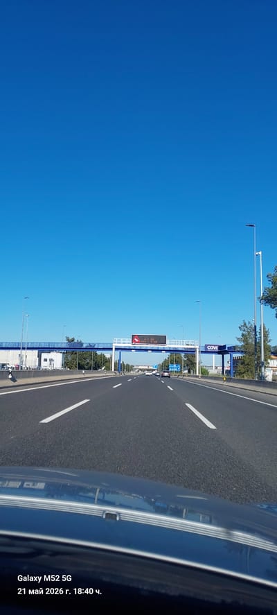 The image shows a view from inside a moving car on a highway near Polígono Industrial De Quart De Poblet, Spain. The sky is clear and blue. The road is paved with white lane markings. Along the side of the road, there are signposts, structures, and trees, including a blue structure labeled "CIVE". A digital billboard is also visible. The edge of the car's exterior is in the right side of the frame.