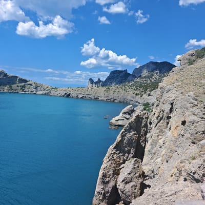 The image shows a rugged coastline with steep, rocky cliffs overlooking a vibrant blue sea under a bright, cloud-dotted sky. In the distance, a series of jagged mountain peaks rise from the land. The foreground is dominated by a rocky, sun-drenched cliff face. The sea is calm with gentle ripples. There are no visible people in the frame. The scene is likely a natural landscape, possibly a hiking trail or viewpoint in a coastal area. The location is identified as the city of Sudak, Ukraine, which is known for its dramatic coastline and mountainous terrain.