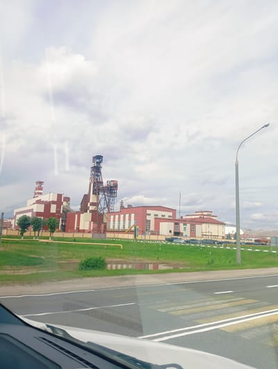 This media file captures a view of an industrial complex situated in Krasnadvorski Sel'ski Savet, Belarus. The scene features a collection of large, functional buildings, primarily colored in red and white, indicating an industrial or manufacturing setting. Dominating the skyline is a tall, intricate metal structure, likely a mining headframe or a similar industrial tower, with visible metal scaffolding and machinery. Adjacent to it are several rectangular buildings, some with multiple stories and numerous windows.
A paved road with a pedestrian crossing is in the foreground, suggesting this view is captured from a vehicle or roadside. The area surrounding the industrial buildings is a grassy landscape, with some trees lining the perimeter and a few yellow pipes visible on the ground. There are several cars parked in front of the buildings, and a lamppost stands tall on the right side of the image. The sky is overcast with white and grey clouds, suggesting a cloudy day. There are no people clearly visible in the image. The overall impression is one of a working industrial area under a subdued sky.