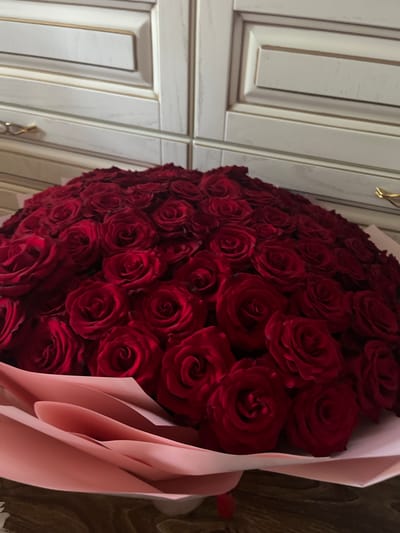 A large bouquet of red roses is displayed in front of cream-colored cabinets with gold accents, suggestive of a kitchen or dining room setting. The flowers are tightly packed and dominate the foreground, with delicate pink paper wrapping peeking out from beneath them. The background shows a portion of ornate kitchen cabinets. The overall impression is one of luxury and a special occasion. This scene is located in Pănășești, Moldova.