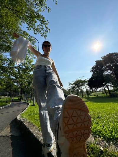 The media shows a low-angle, wide shot of a young woman in a park on a sunny day. The sun is bright and high in the sky, indicating it is likely midday. The woman is wearing a white tank top and light-wash denim jeans. She is smiling and holding a white piece of fabric, possibly a dress or skirt, which billows out to her right. Her left hand is extended, holding the fabric. A black handle, likely from a bag or basket, is visible. Her right foot, wearing a light-colored sneaker, is prominently featured in the foreground, with its patterned sole facing the camera.

The setting is a park with lush green grass and trees. The background shows more trees and a slightly blurred landscape, suggesting a natural or recreational area. The overall mood appears cheerful and relaxed, conveyed by the woman's smile and the bright, sunny weather. The location is identified as Thuận Nam, Vietnam. There is no visible text in the image.