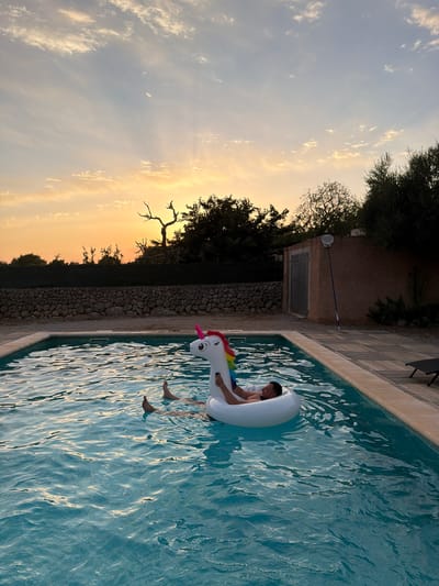 A man is relaxing on an inflatable unicorn in a swimming pool at sunset. The pool is surrounded by a paved patio and a stone wall. In the background, there are trees and a building. The sky is orange and yellow, with some clouds. The scene appears to be in a rural area, possibly a villa or resort, in the municipality of Santa Margalida, Spain.