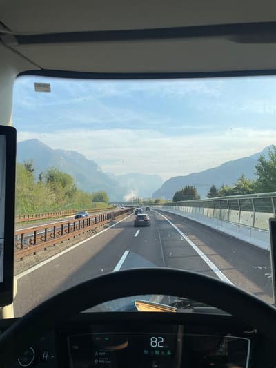 The image is taken from inside a vehicle, possibly a truck, driving on a highway in Rivalta, Italy. The camera is positioned behind the steering wheel, showing a view through the windshield. On the right, the vehicle's speedometer indicates a speed of 82 km/h. Outside, the highway has multiple lanes with a safety barrier along the side. A railway track runs parallel to the highway on the left side. Further in the distance, mountains are visible under a clear, somewhat hazy sky. Other vehicles are present on the road ahead. The speed limit sticker on the windshield indicates 90 km/h, 56 mph.