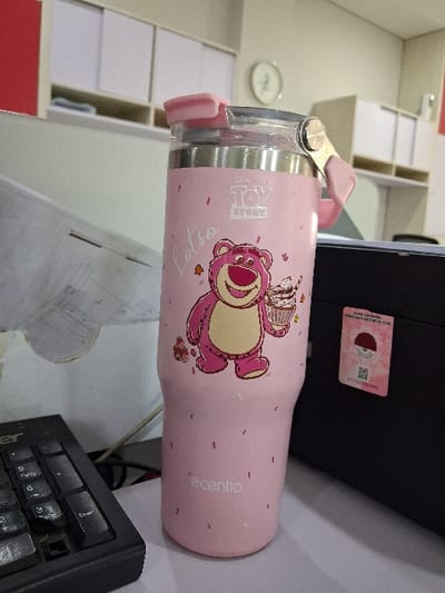 A pink tumbler with a Toy Story Lotso bear design, holding a cupcake, sits near a computer. The tumbler has a clear lid with pink accents and the brand "ecentio" printed on it. A black computer tower and a keyboard are visible in the background. The scene includes a desk with some papers and a cabinet with shelves.