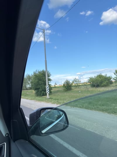 The image captures a view from inside a car traveling down a road. The scene is set outdoors under a bright blue sky with scattered white clouds, suggesting a clear day. The time of day appears to be daytime, likely late morning or early afternoon, given the strong sunlight.
The prominent subjects include the car's side mirror, reflecting the sky and trees, and the road with a white lane marking. Beyond the road, there is a grassy area with several trees and a tall pole with a black and white striped marker. The landscape suggests a rural or semi-rural setting. The overall mood is calm and ordinary, depicting a typical journey.
The image is oriented vertically. Visible text is not present in the image. The location context is Novaya Usman, Russia.