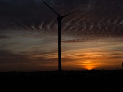 This media file captures a dramatic sunset or sunrise over a flat landscape, featuring a large wind turbine silhouetted against the colorful sky.
The scene is dominated by the striking silhouette of a wind turbine positioned slightly to the left of the center. Its tall, slender mast extends vertically, with two of its three blades visible, angled upwards to form a 'V' shape against the sky. The entire wind turbine is cast in deep shadow, rendering it black.
The sky is a prominent feature, showcasing a gradient of colors characteristic of golden hour. The upper portion of the sky is a dark, moody gray-purple with distinct, wavy cloud formations, possibly indicating altocumulus or mammatus clouds. These textured clouds transition downwards into warmer hues, giving way to soft oranges, yellows, and faint reds closer to the horizon. The sun, a brilliant semi-circle of intense orange and yellow light, is visible just above the horizon line on the right side of the frame, partially obscured by the distant landscape.
The horizon itself is a dark, indistinct silhouette of what appears to be an industrial area or distant town, with scattered structures, possibly buildings or smaller communication towers, and a low, flat tree line. The foreground is entirely black, suggesting open land or water also in deep shadow, further emphasizing the vastness of the sky.
The image conveys a peaceful yet powerful atmosphere, highlighting the beauty of nature and industrial elements coexisting. There is no visible text or identifiable human activity. Given the location context of Maasdijk, Netherlands, the scene aligns with the flat, often windy landscapes where wind energy is common.