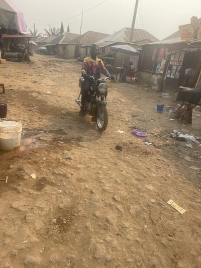 This image depicts an unpaved street in Dzhidu, Nigeria, characterized by a dusty, hazy atmosphere.
In the mid-ground, a young adult male with dark skin is seated on a green motorcycle, facing directly towards the viewer with a slight smile. He wears a vibrant, multi-colored long-sleeved shirt patterned with what appear to be multiple faces, and dark trousers. A black bag is secured behind the motorcycle's seat. The motorcycle features a single round headlight and a visible chrome exhaust pipe on its right side.
The ground consists of uneven, light-brown packed dirt with visible rocks, ruts, and scattered debris including plastic bags, wrappers, and other refuse. To the immediate left foreground, a white plastic bucket containing liquid or residue is visible, surrounded by a damp patch on the ground. Further back on the left, a makeshift stall with a light-colored fabric awning stands.
On the right side of the street, multiple single-story buildings with corrugated metal roofs line the road. These appear to be small shops or businesses, with one sign partially legible as "ZYENG ENTERPRISE" with "POS" below it. Another individual is partially visible seated under a light-colored umbrella or awning in front of one of these shops. A dark blue bucket and additional scattered items are on the ground near these structures.
The background shows the dirt road continuing, flanked by more similar buildings and sparse vegetation, including palm trees on the left. The sky is hazy, indicating a dry or dusty environment, and casts a diffused, bright light over the scene.
